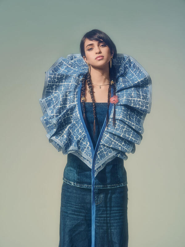 Girl in jeans dress wearing a blue floral patterned ruffled sash over her shoulders.