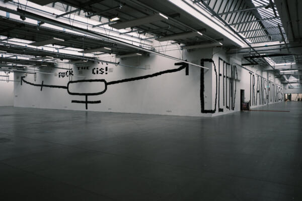 En gigantisk mural av svart text och symboler på vit bakgrund fyller två vägg runt ett hörn i ett stort öppet utrymme. Texten säger "Fuck the Cis!" och "Defend Trans Futures!"